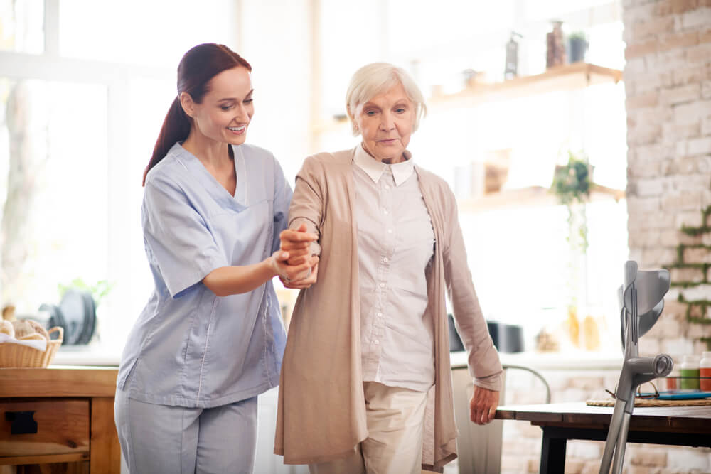 Aged Grey-Haired Woman Walking After Surgery With the Help of Nurse