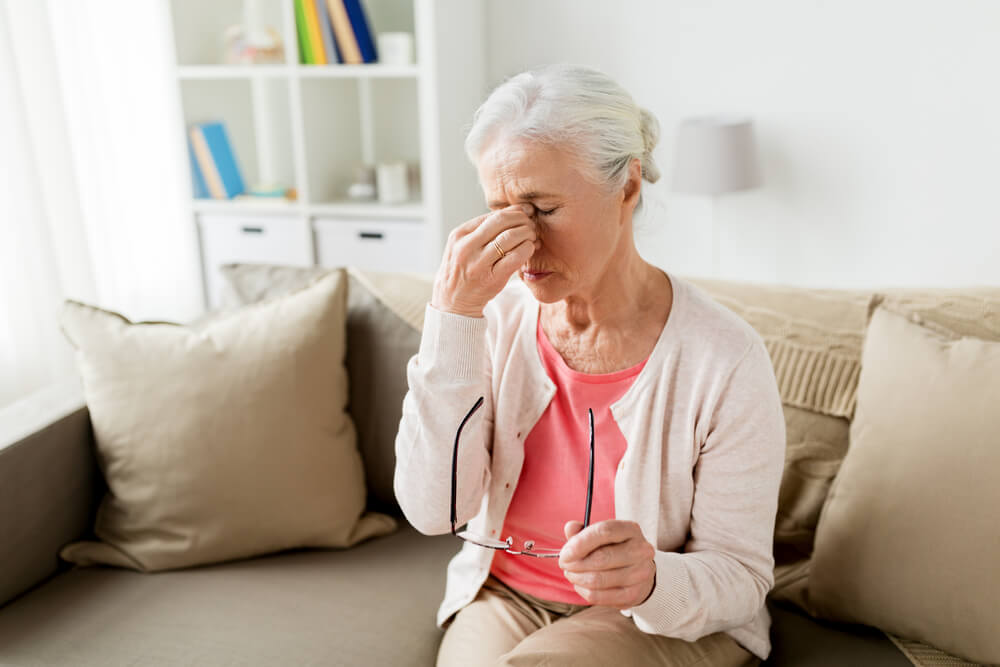 Senior Woman With Glasses Sitting on Sofa and Having Headache at Home